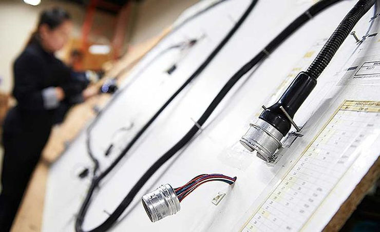 Worker assembling complex wiring on a large board in a workshop; focus on multicolored wires and connectors, blurred background.